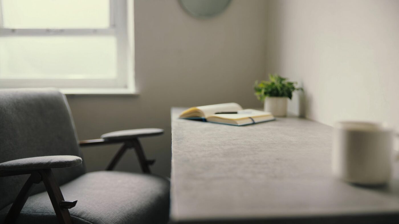 A quiet consulting room desk beside a window, with a notebook, pen, plant and mug in soft daylight.