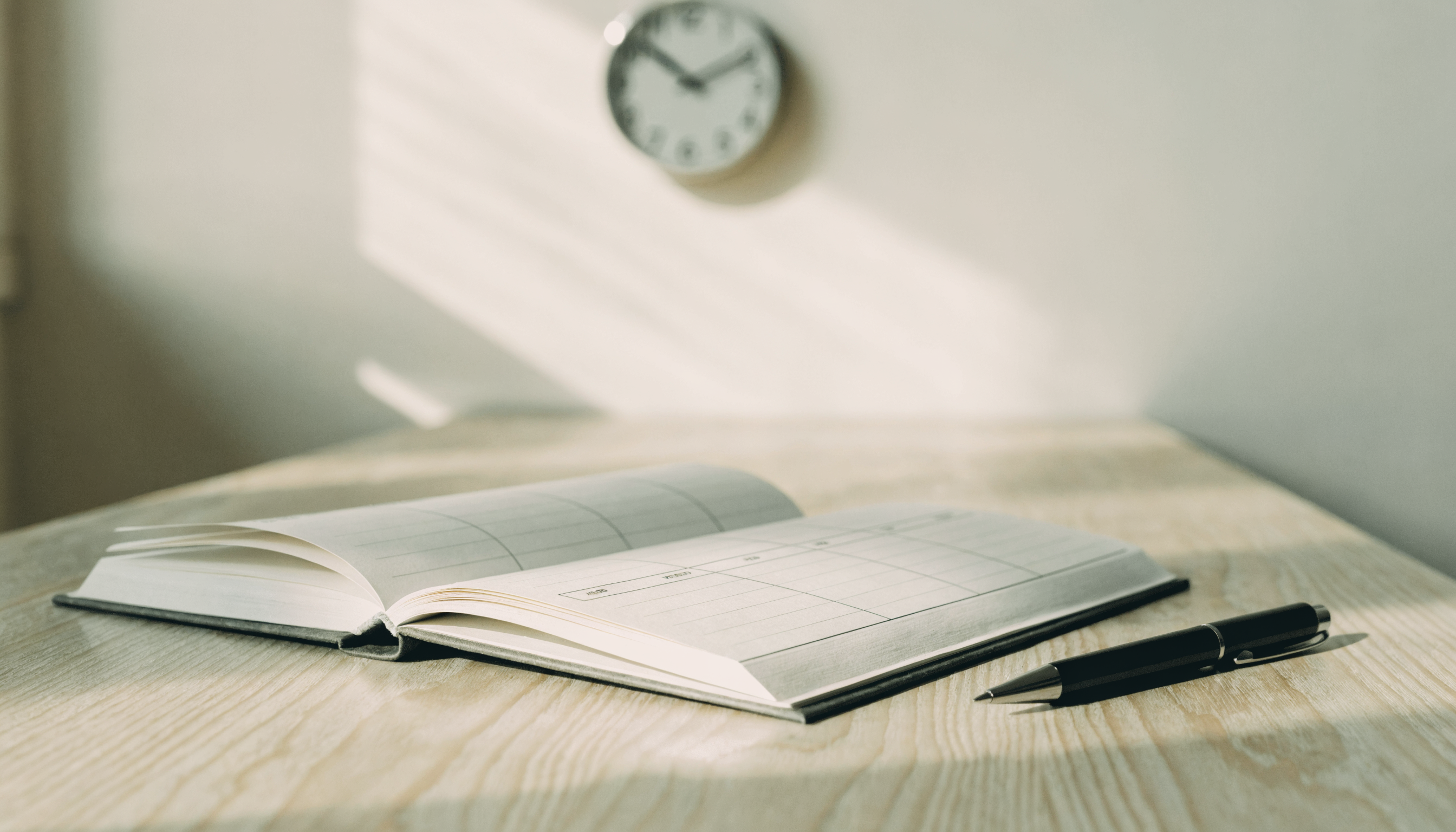Open paper planner and pen on a dry sunlit table, soft wall clock in the background