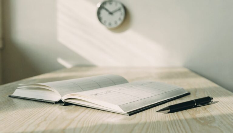 Open paper planner and pen on a dry sunlit table, soft wall clock in the background